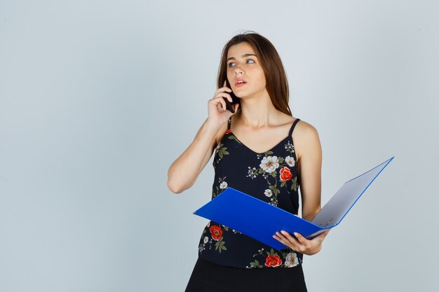 Portrait of young lady holding folder, talking on mobile phone in blouse and looking pensive front view