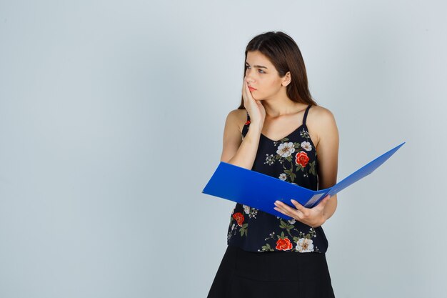 Portrait of young lady holding folder while keeping hand on cheek in blouse, skirt and looking thoughtful front view