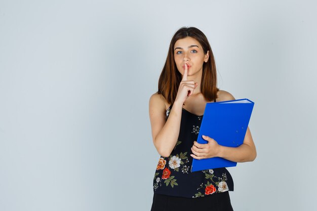 Portrait of young lady holding folder while showing silence gesture in blouse, skirt and looking careful front view