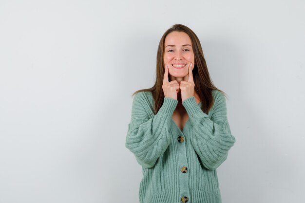 Portrait of young lady keeping fingers on cheeks in wool cardigan and looking glad front view