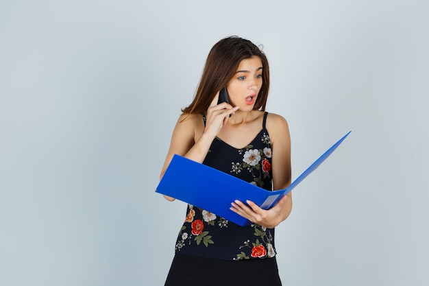 Portrait of young lady looking through folder while talking on mobile phone in blouse and looking shocked front view