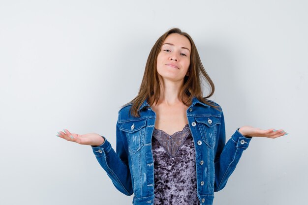 Portrait of young lady making scales gesture in blouse, denim jacket and looking cheerful front view