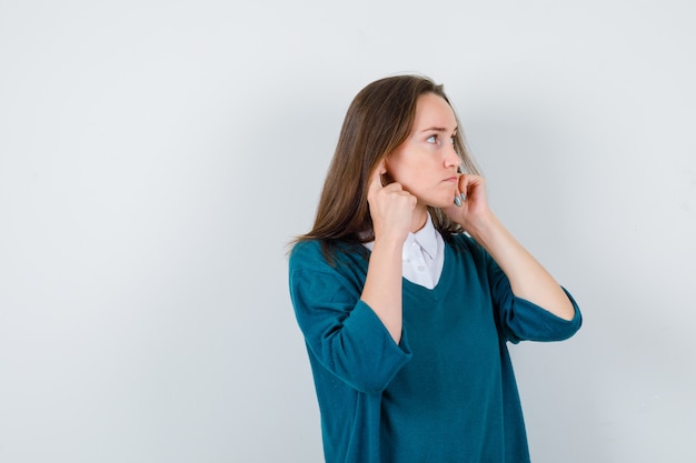 Portrait of young lady plugging ears with fingers, looking aside in white shirt, sweater and looking focused front view