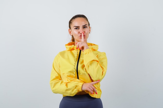 Portrait of young lady pointing to the right side, showing silence gesture in yellow jacket and looking confident front view