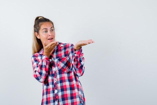 Portrait of young lady showing something in checked shirt and looking amazed front view