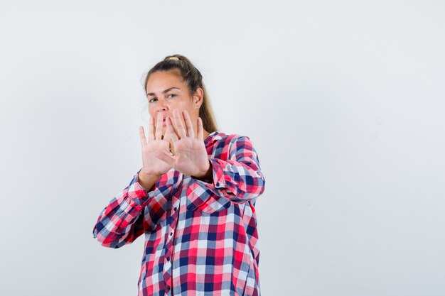 Portrait of young lady showing stop gesture in checked shirt and looking scared front view