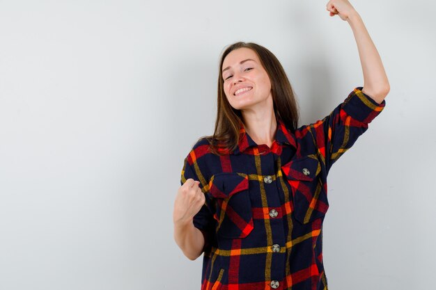 Portrait of young lady showing winner gesture in casual shirt and looking blissful front view