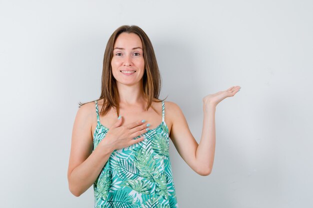 Portrait of young lady with hand on chest while holding something in blouse and looking cheery front view