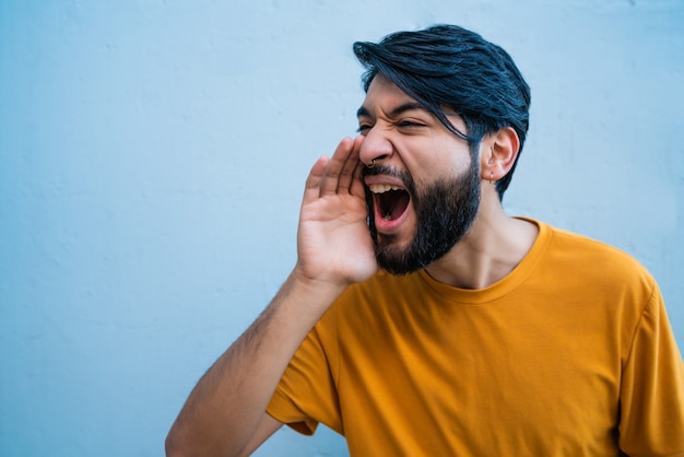 Portrait of young latin man shouting and screaming against blue backgound.