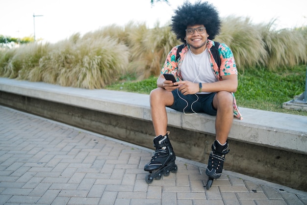 Portrait of young latin man using his mobile phone and wearing skate rollers while sitting outdoors. Sports and urban concept