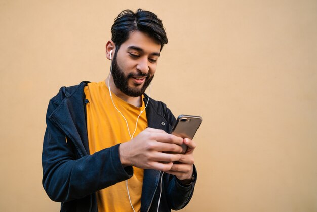 Portrait of young latin man using his mobile phone with earphones against yellow space. Communication concept.