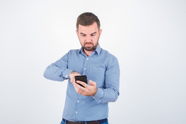 Portrait of young male looking at mobile phone in shirt, jeans and looking focused front view