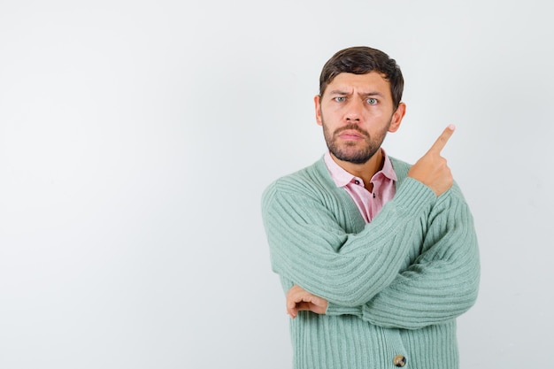 Free photo portrait of young male pointing at upper right corner in shirt, cardigan and looking hesitant front view