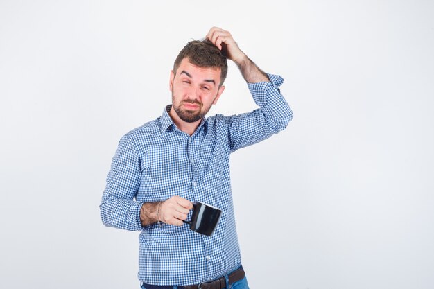 Portrait of young male scratching head while holding cup in shirt, jeans and looking pensive front view