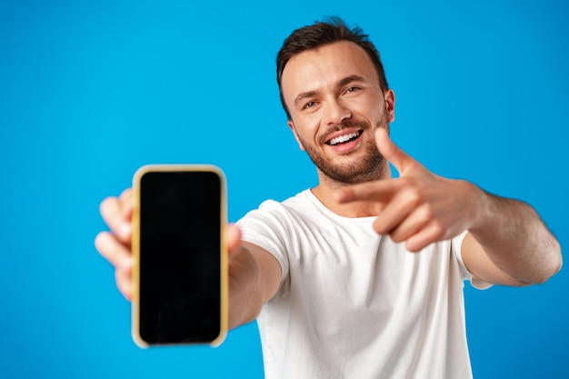 Portrait of young man advertising new smartphone showing it to camera against blue background