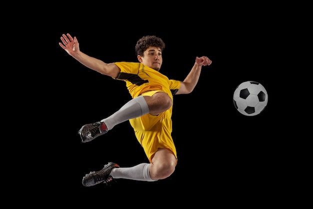 Free photo portrait of young man football playing in motion training isolated over black studio background kicking ball