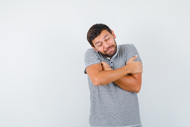 Portrait of young man hugging himself, keeping eyes shut in t-shirt and looking meek front view