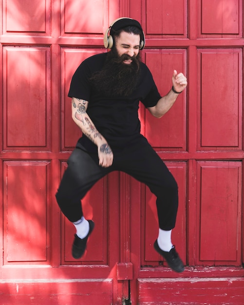 Free photo portrait of a young man jumping in air with headphone on his ear against red door