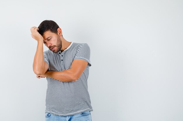 Portrait of young man keeping hand on head bent down in striped t-shirt and looking depressed front view