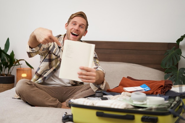 Free photo portrait of young man pointing at his list of items packing clothes on vacation showing notebook