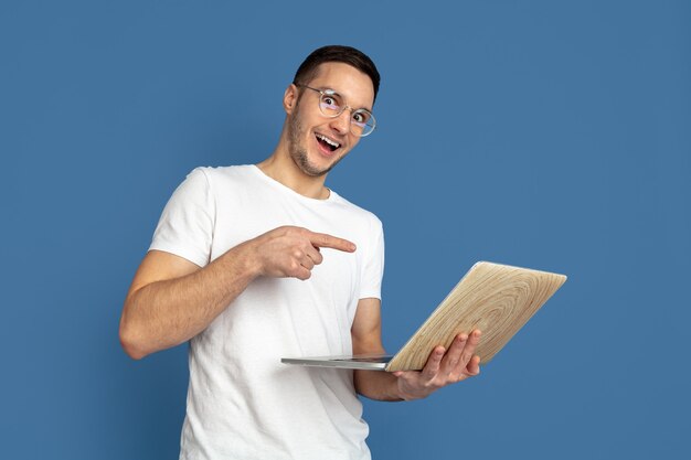 Portrait of young man pointing to laptop isolated on blue studio wall