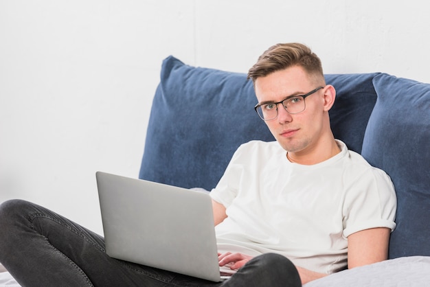 Free photo portrait of a young man sitting on bed with laptop looking at camera