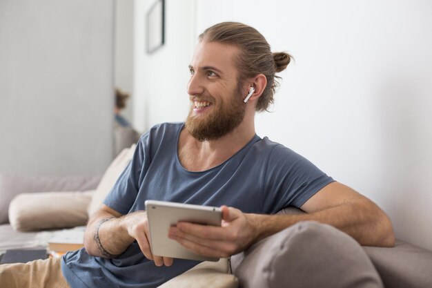 Portrait of young man sitting on gray sofa with earphones and the tablet in hands and happily looking aside at home