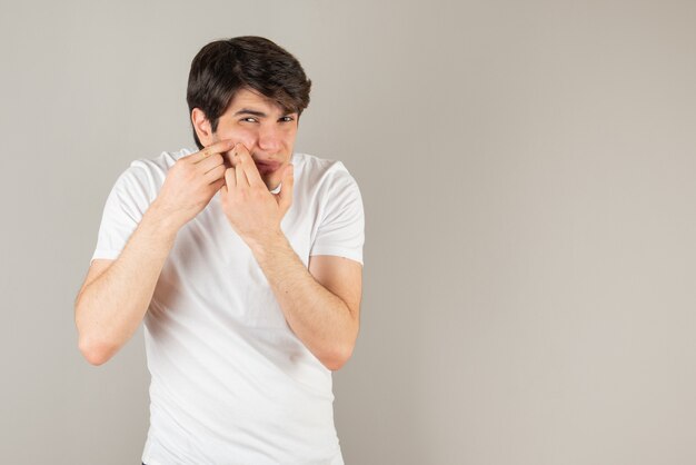 Portrait of young man squeezing his pimple against gray.