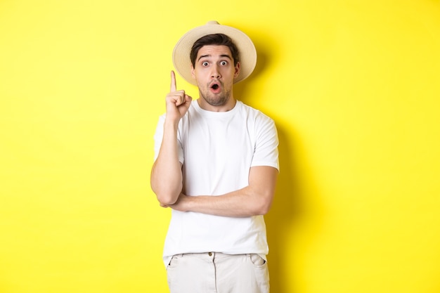 Portrait of young man in straw hat having an idea, raising finger eureka sign, making suggestion, standing over yellow background.