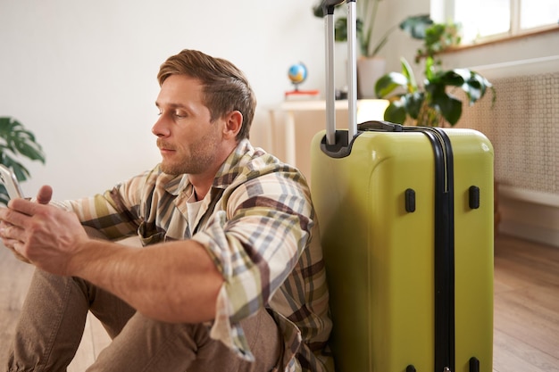 Free photo portrait of young man traveller sits with suitcase and mobile phone chats on smartphone