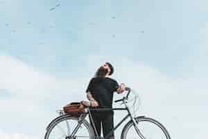 Free photo portrait of a young man with bag on his bicycle looking at birds flying in the sky