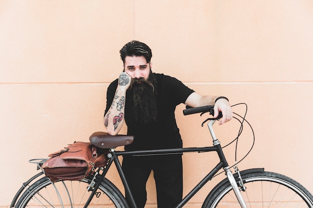 Free photo portrait of a young man with tattoo on his hand standing with bicycle against wall