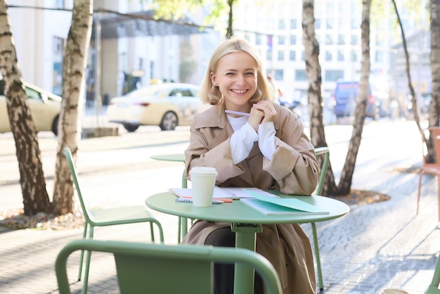 Free photo portrait of young modern woman in stylish trench coat sitting in outdoor cafe on sunny day writing