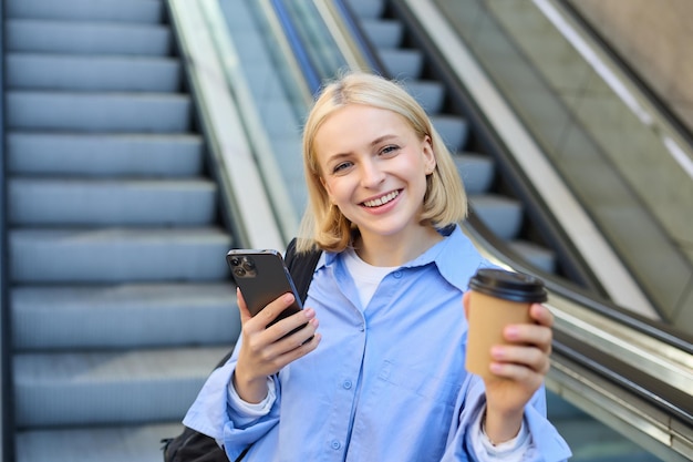 Free photo portrait of young modern woman with backpack smartphone drinking cup of coffee standing near