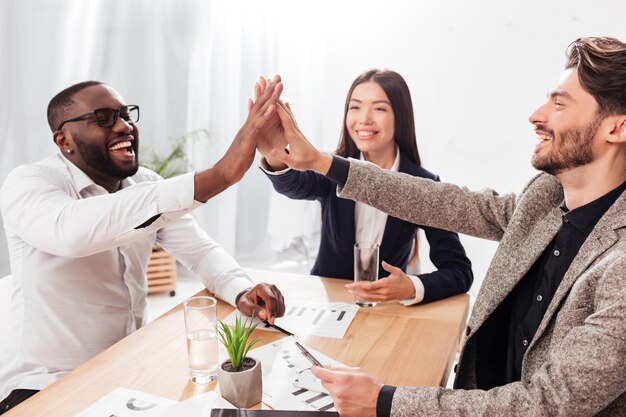 Portrait of young multinational group of business partners sitting at the table and joyfully giving high five to each other while working together in office isolated