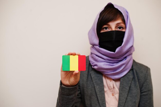 Portrait of young muslim woman wearing formal wear protect face mask and hijab head scarf hold Mali flag card against isolated background Coronavirus country concept