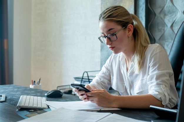 Portrait of young office worker woman sitting at office desk with documents holding smartphone looking at screen with serious expression on face working in office