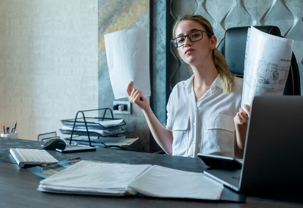 Portrait of young office worker woman sitting at office desk with documents looking at camera with serious confident expression on face working in office