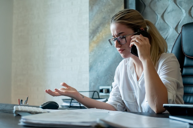 Portrait of young office worker woman sitting at office desk with documents talking on mobile phone nervous and stressed working in office