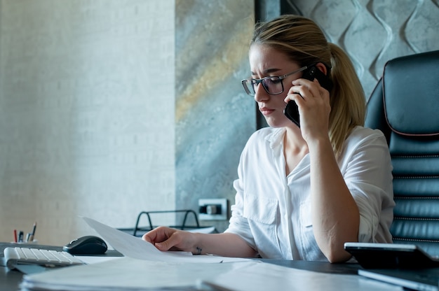 Portrait of young office worker woman sitting at office desk with documents talking on mobile phone nervous and stressed working in office