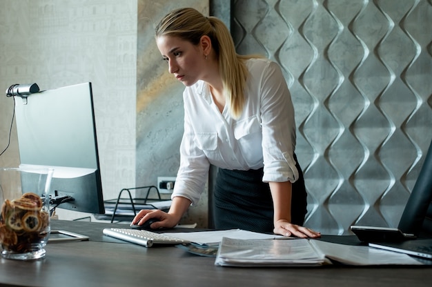 Free photo portrait of young office worker woman standing at office desk with documents using computer with confident and serious expression on face working in office