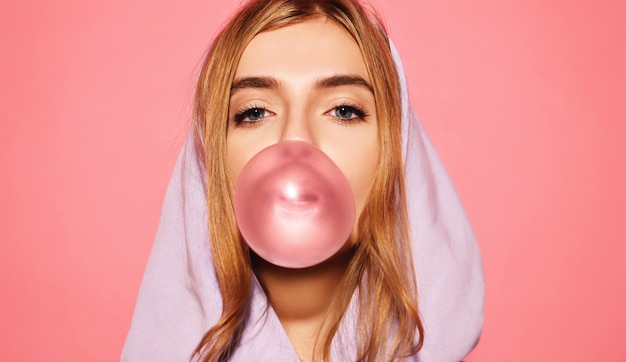 Free photo portrait of young playful blonde women dressed in blue hoodie standing isolated over pink wall while blowing bubble with chewing gum. women