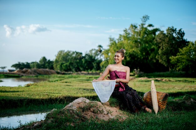 Portrait Young pretty Asian woman in beautiful Thai traditional clothes at rice field, she sitting near fishing equipment