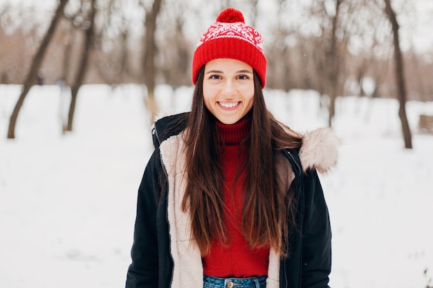 Portrait of young pretty smiling happy woman in red sweater and knitted hat wearing winter coat, walking in park in snow, warm clothes