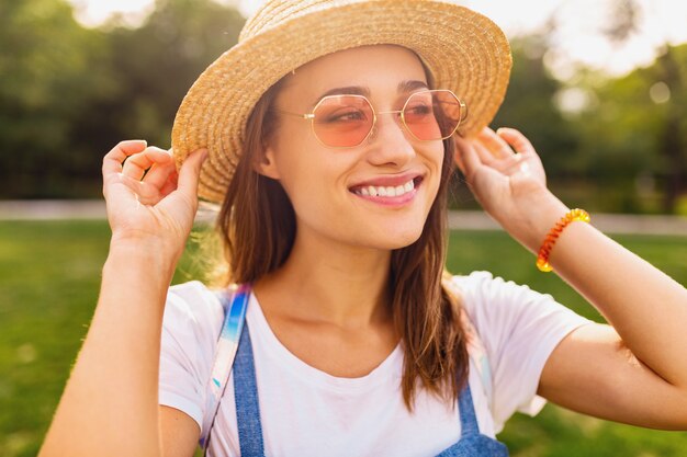 Portrait of young pretty smiling woman in straw hat and pink sunglasses walking in park, summer fashion style, colorful hipster outfit