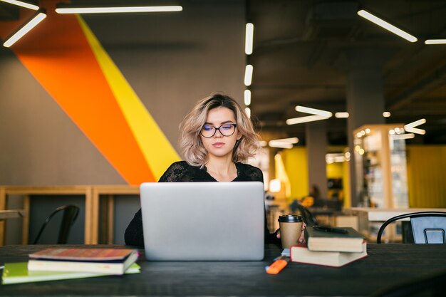 Portrait of young pretty woman sitting at table in black shirt working on laptop in co-working office