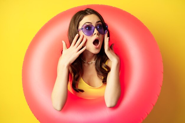 Portrait of young pretty woman in sunglasses, wearing beach swimming ring, looking surprised and amazed, shocked wow face, yellow background