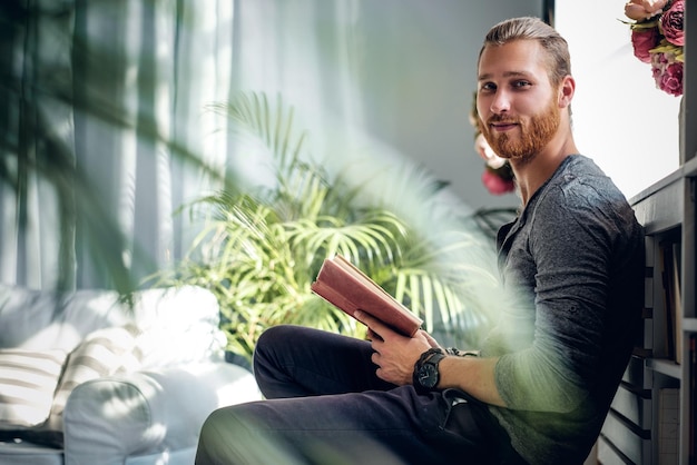 Free photo portrait of young redhead bearded male holding a book in a room with green plants.