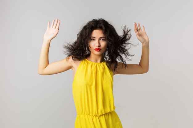 Free photo portrait of young sexy brunette woman in yellow dress, curly hair waving, summer style, fashion trend, , mixed race, isolated, holding hands up