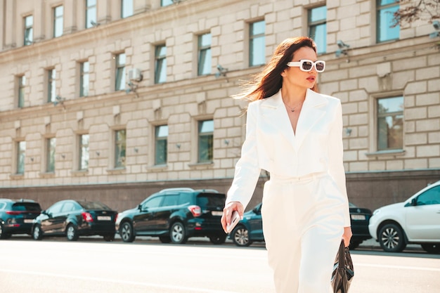 Portrait of young sexy modern businesswoman model. Hot beautiful woman in white suit posing on the street background. Fashion female walking outdoors in sunglasses